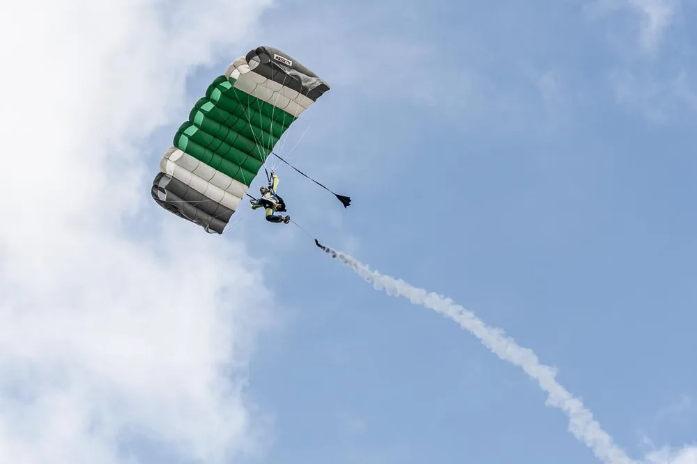 A skydiver trails smoke beneath a green and white parachute high above the park.