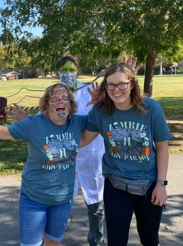 Two smiling women in Zombie Bike Parade shirts pose with a costumed zombie behind them.