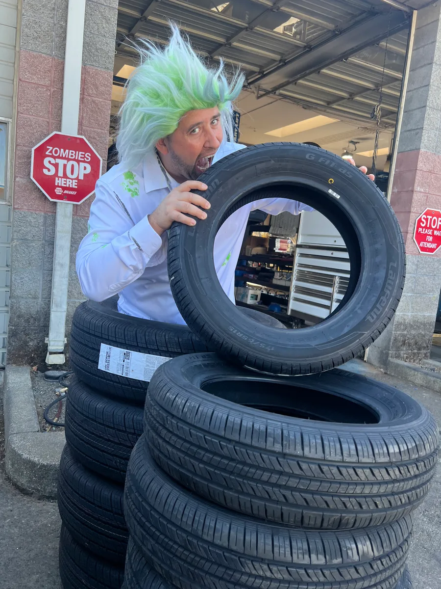 A man in a green zombie wig peers through the middle of a car tyre beside a stack of tyres, next to a red sign reading Zombies Stop Here.