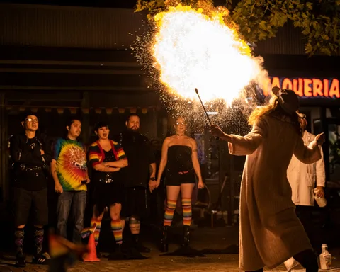 A fire performer sends a burst of flame into the night as a crowd watches on a downtown sidewalk.