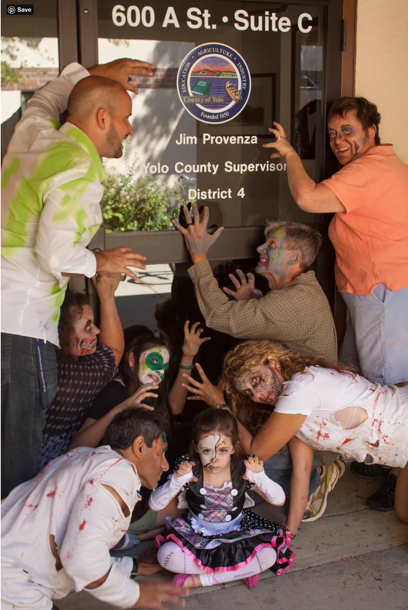 A costumed zombie reaches through an office window as a staff member laughs.