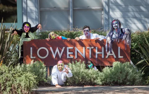 Costumed zombies peek and crawl over a business sign reading Lowenthal.