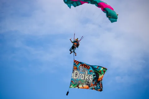 A skydiver gives a thumbs up beneath a colorful parachute towing a banner for The Drake.