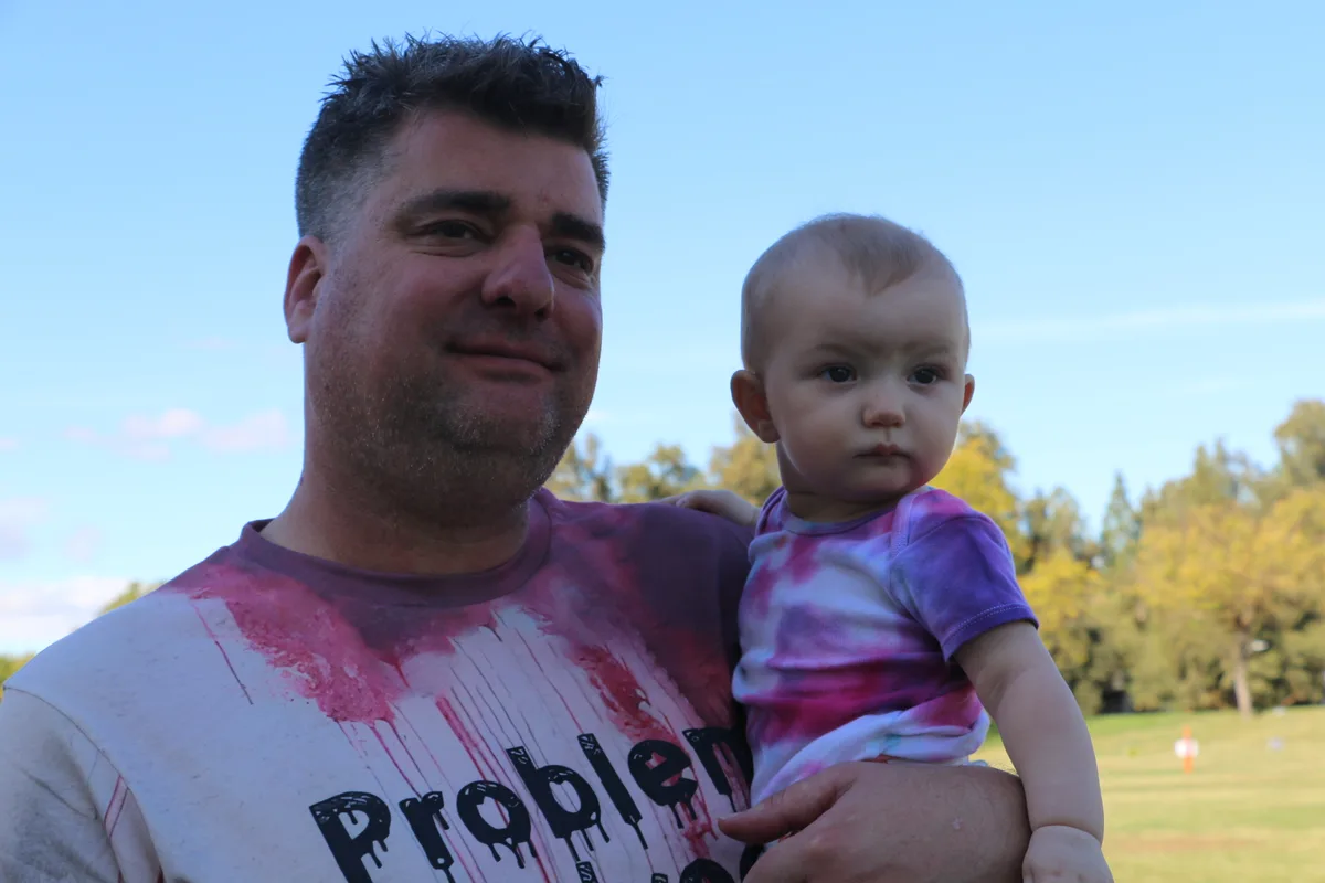 A father in a tie-dye shirt holds his baby in the park during the festival.
