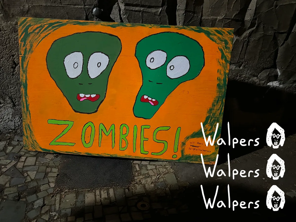 A hand painted wooden sign reading Zombies with two green monster faces, by Will Alpers.