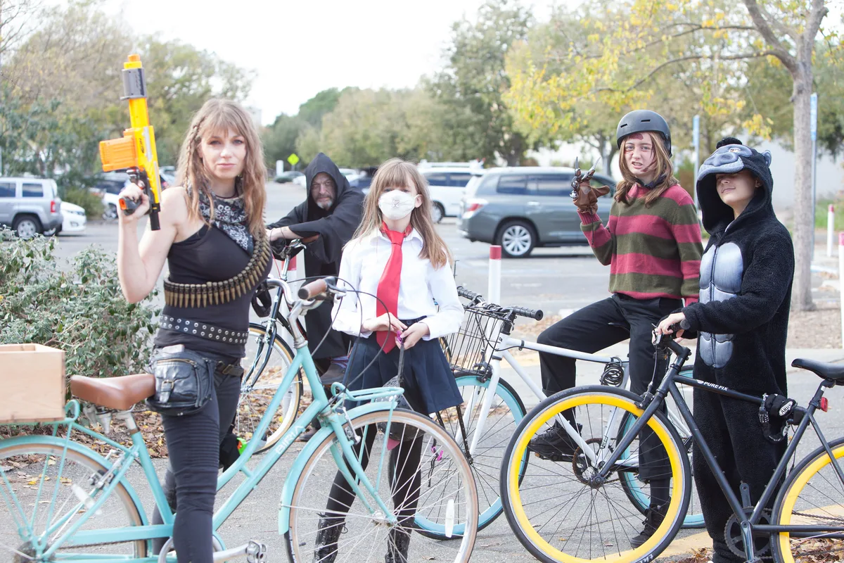 Three women in costume pose with their bikes on a residential street.