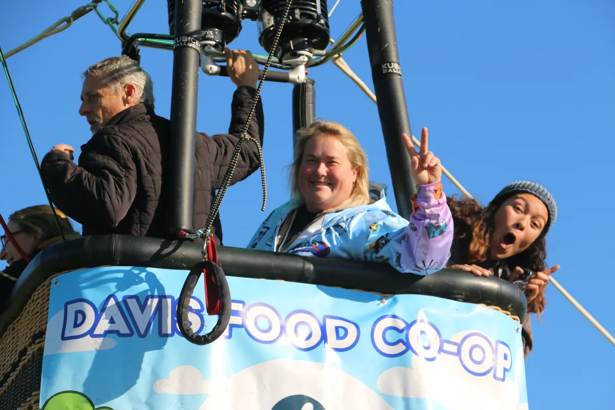 Three riders smile and wave from inside a Davis Food Co-op hot air balloon basket.