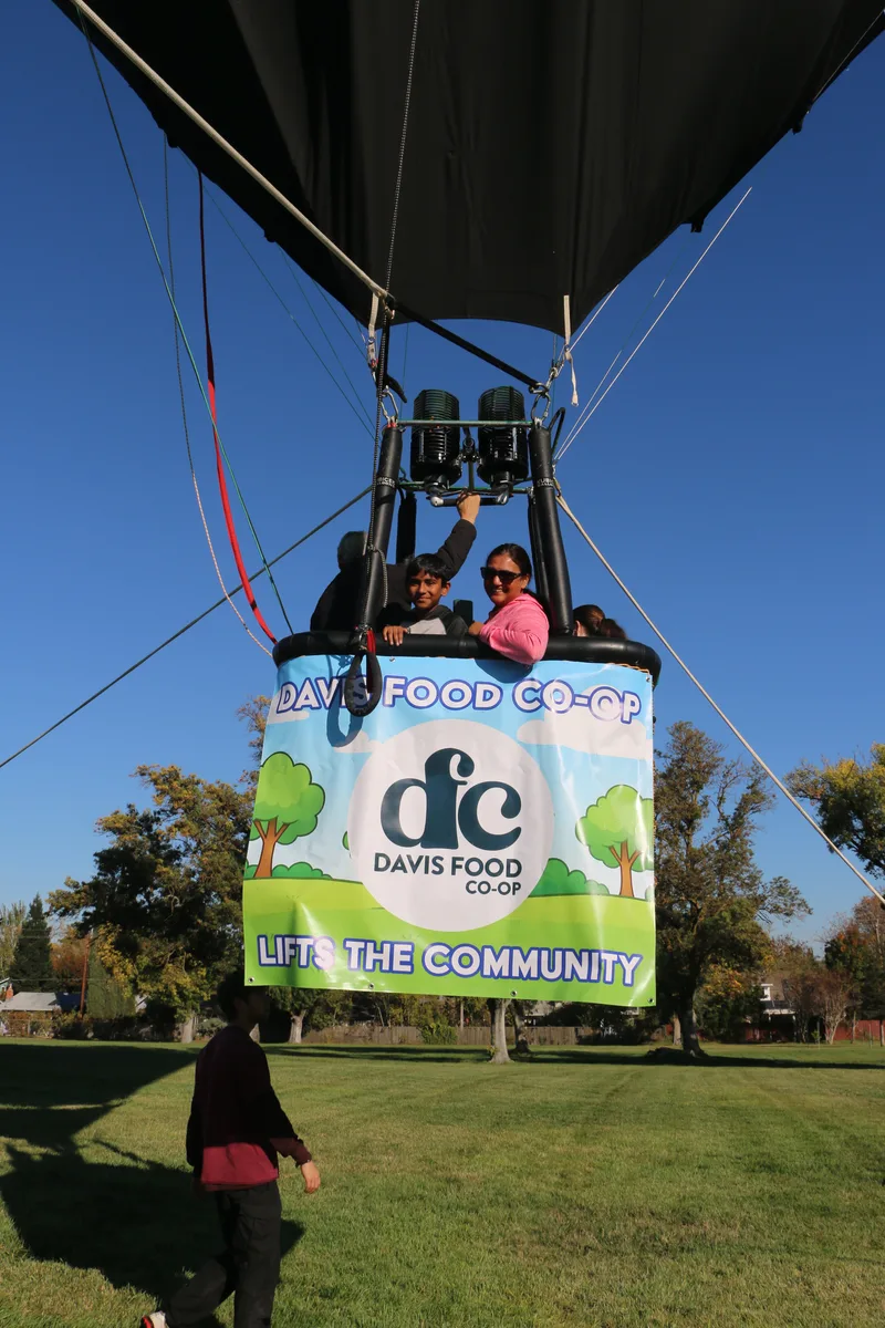 People wave from a hot air balloon basket decorated with a Davis Food Co-op banner.