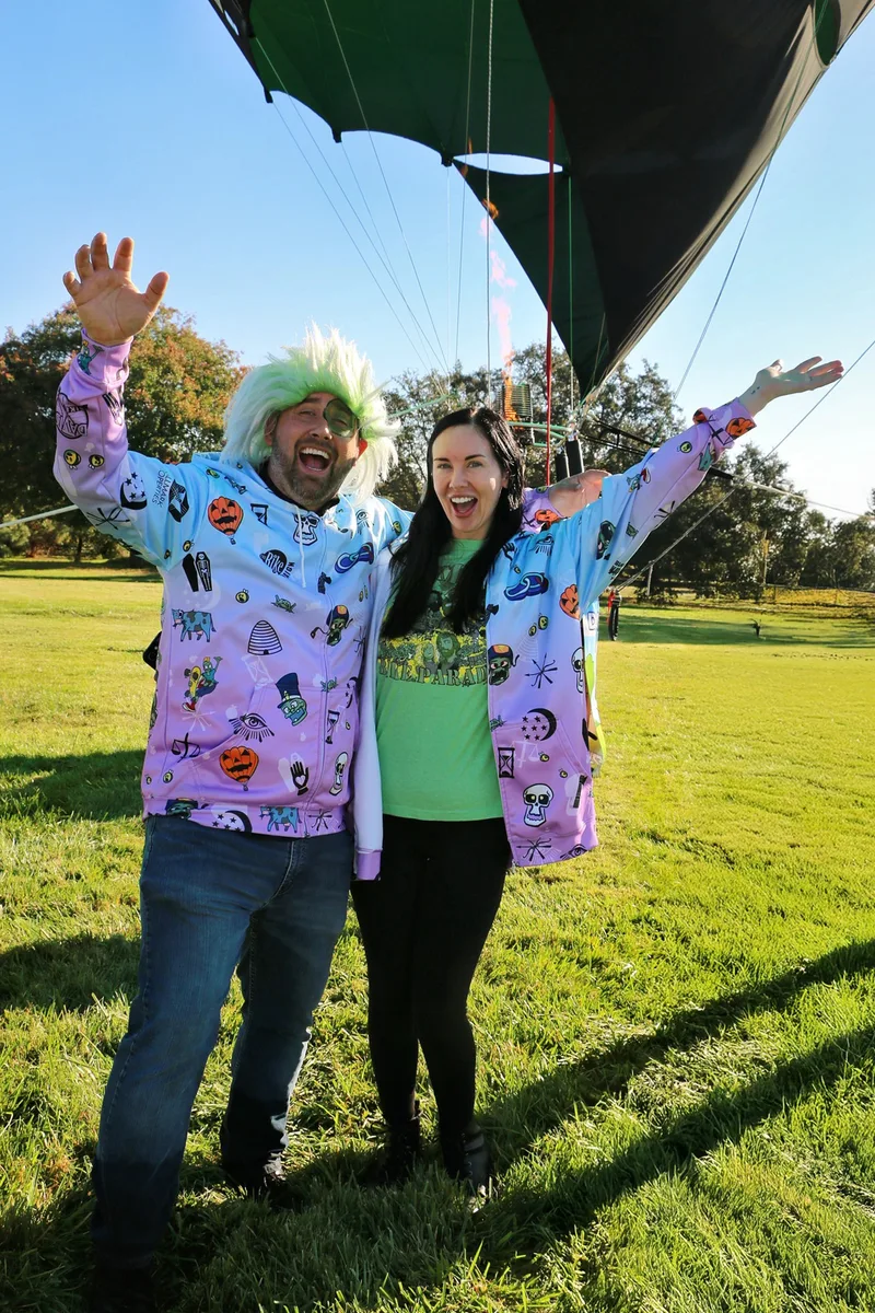 A couple in matching Zombie Bike Parade jackets throw their arms up laughing beside the hot air balloon.