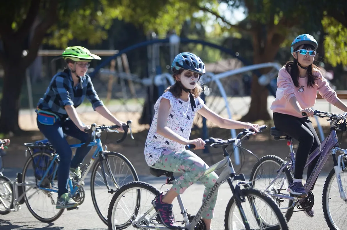 Two kids with skull face paint ride their bikes together in the park.