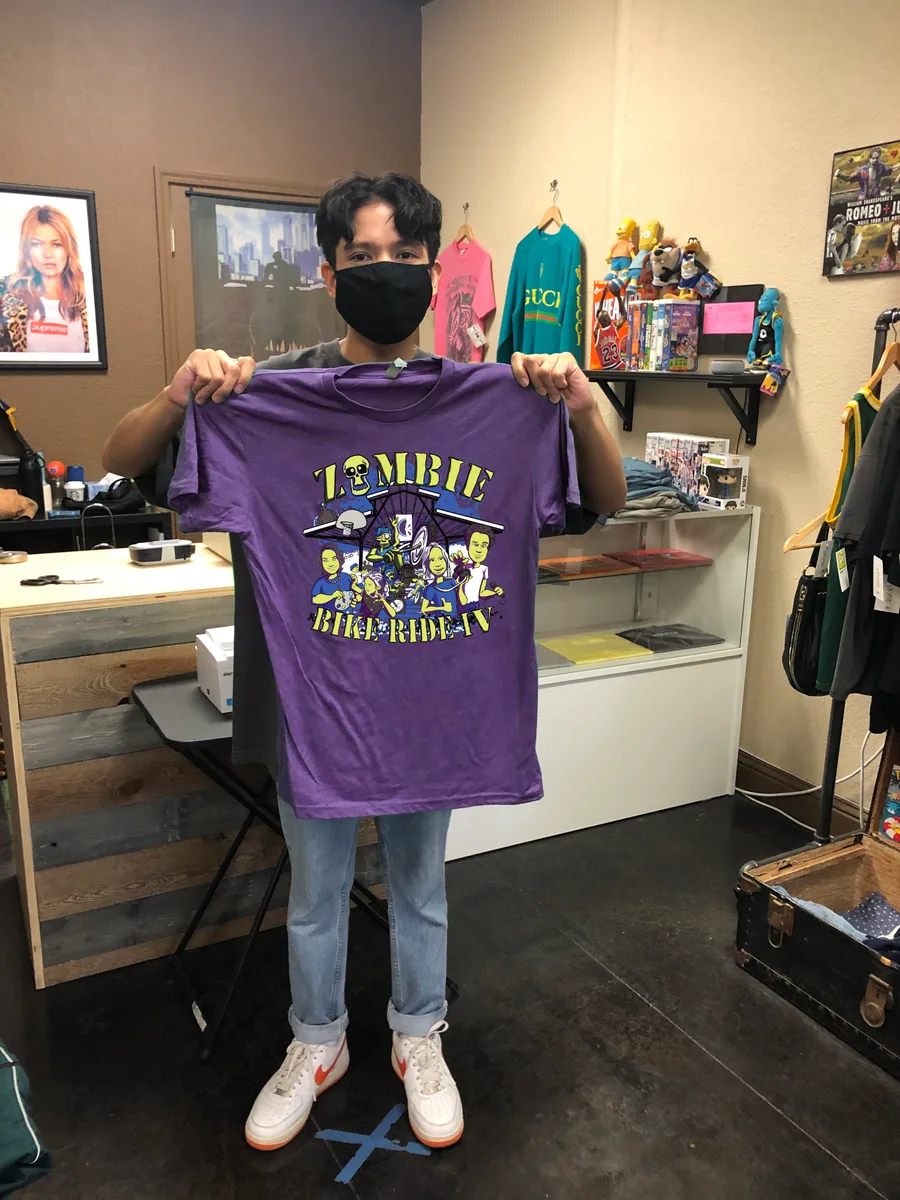 A man in a mask holds up a purple Zombie Bike Ride t-shirt in a shop.