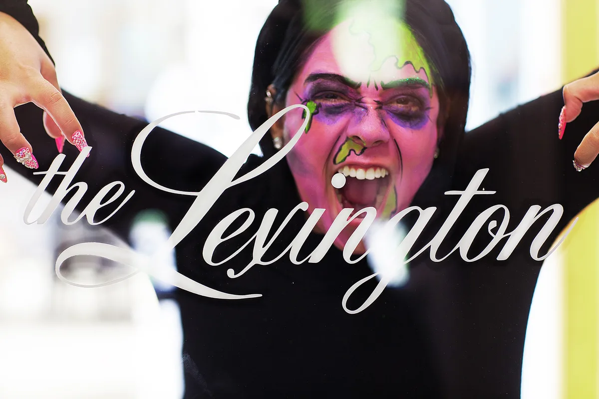 A reveller in green and purple zombie face paint grins with her tongue out and arms raised, under the script logo of The Lexington.