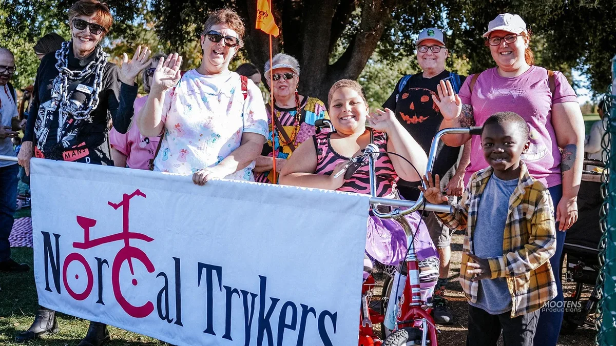 A group of adults and a young boy hold a NorCal Trykers banner and wave beside a decorated adaptive trike.