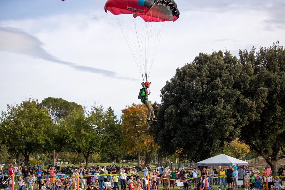 A skydiver descends over the park as a large crowd watches from behind a taped-off landing area.