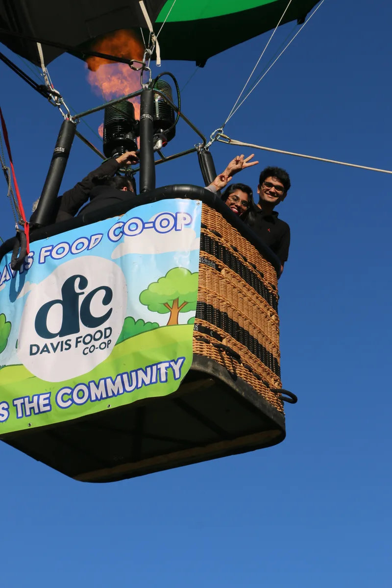 Riders lean out of a hot air balloon basket branded with the Davis Food Co-op logo.