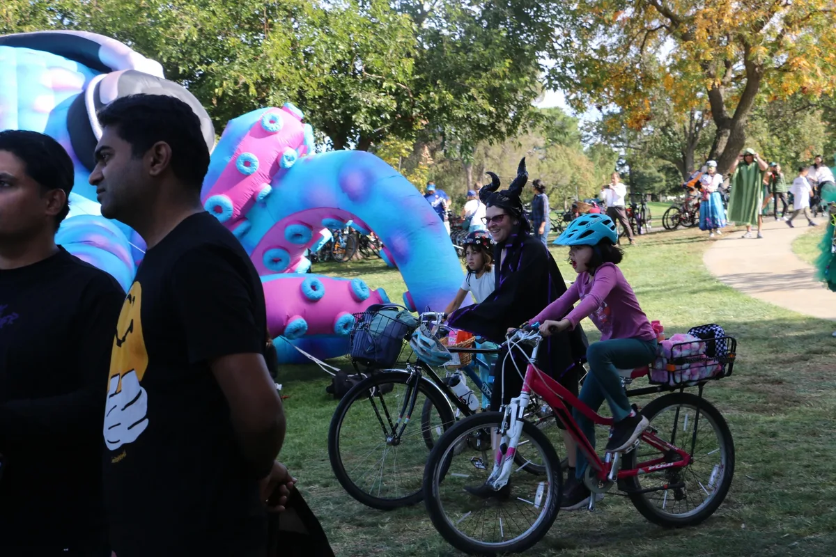 Riders walk their bikes past giant inflatable costume sculptures in the park.
