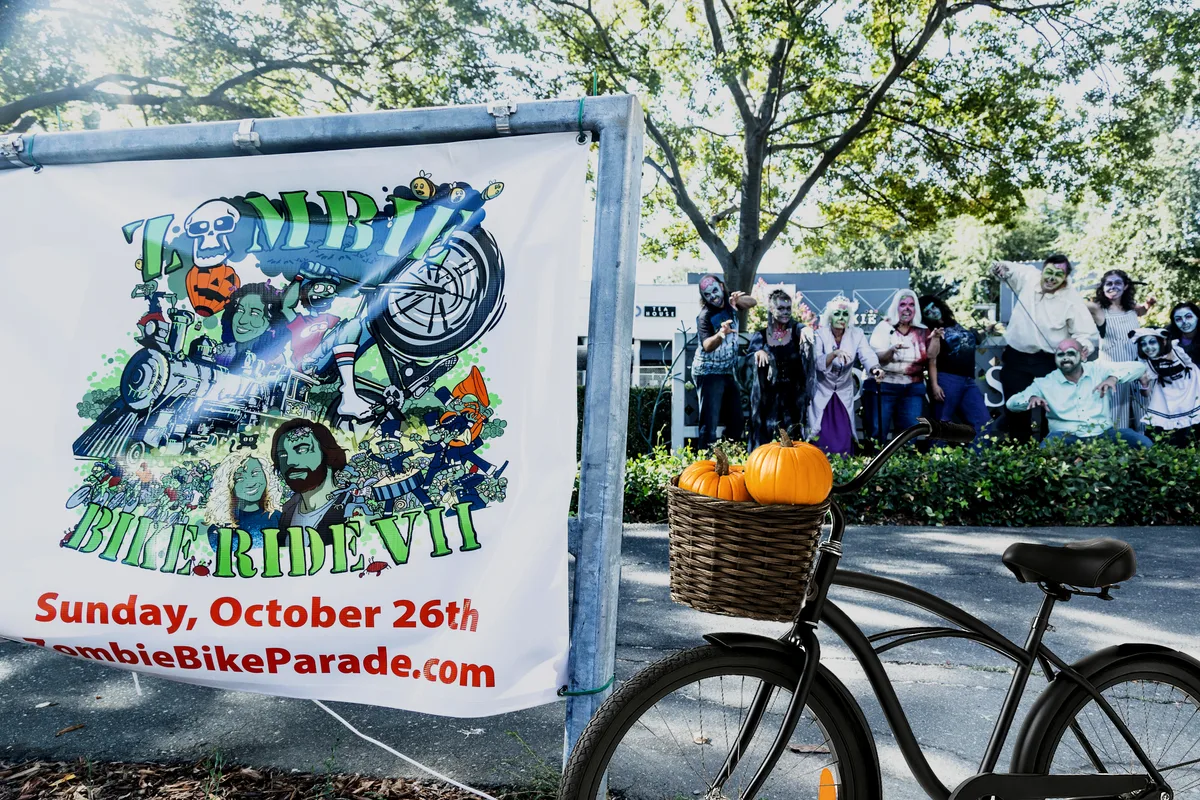 A Zombie Bike Ride banner hangs near a bike and pumpkins with a crowd gathering behind.