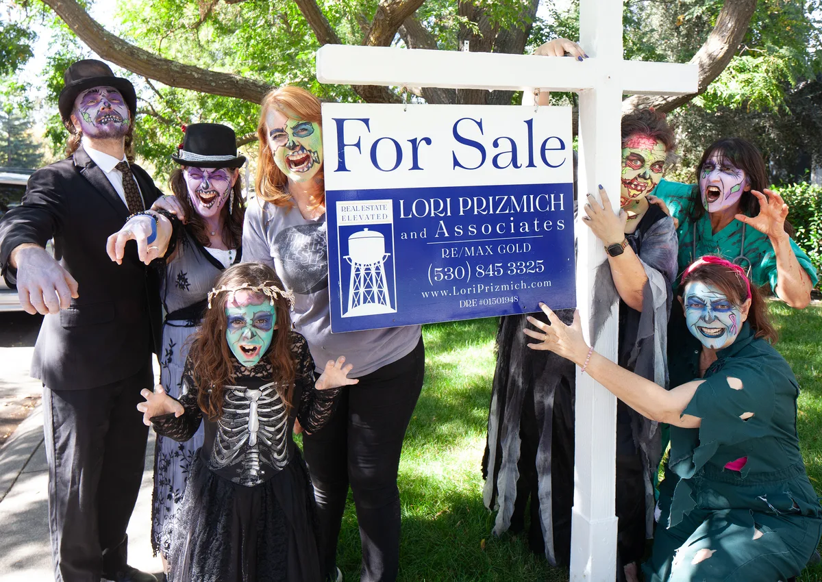 Three people in zombie makeup pose beside a For Sale real estate sign.