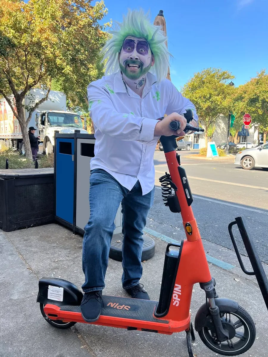 A man in a green wig and lab coat costume smiles on a downtown street.