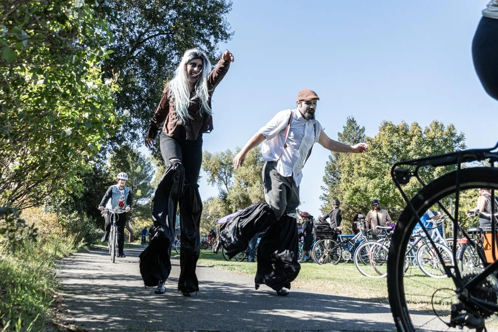 A stilt walker in a flowing black costume strides down a sunny park path with a companion.
