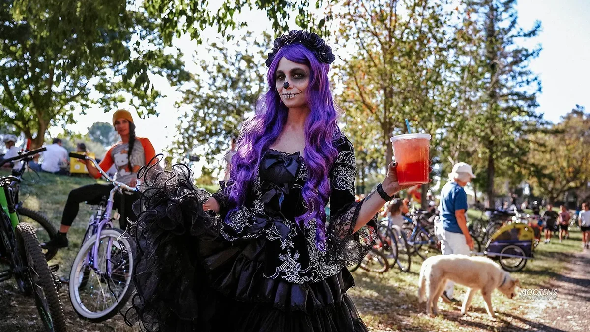 A woman in a purple wig and gothic black dress walks her bike along the park path.