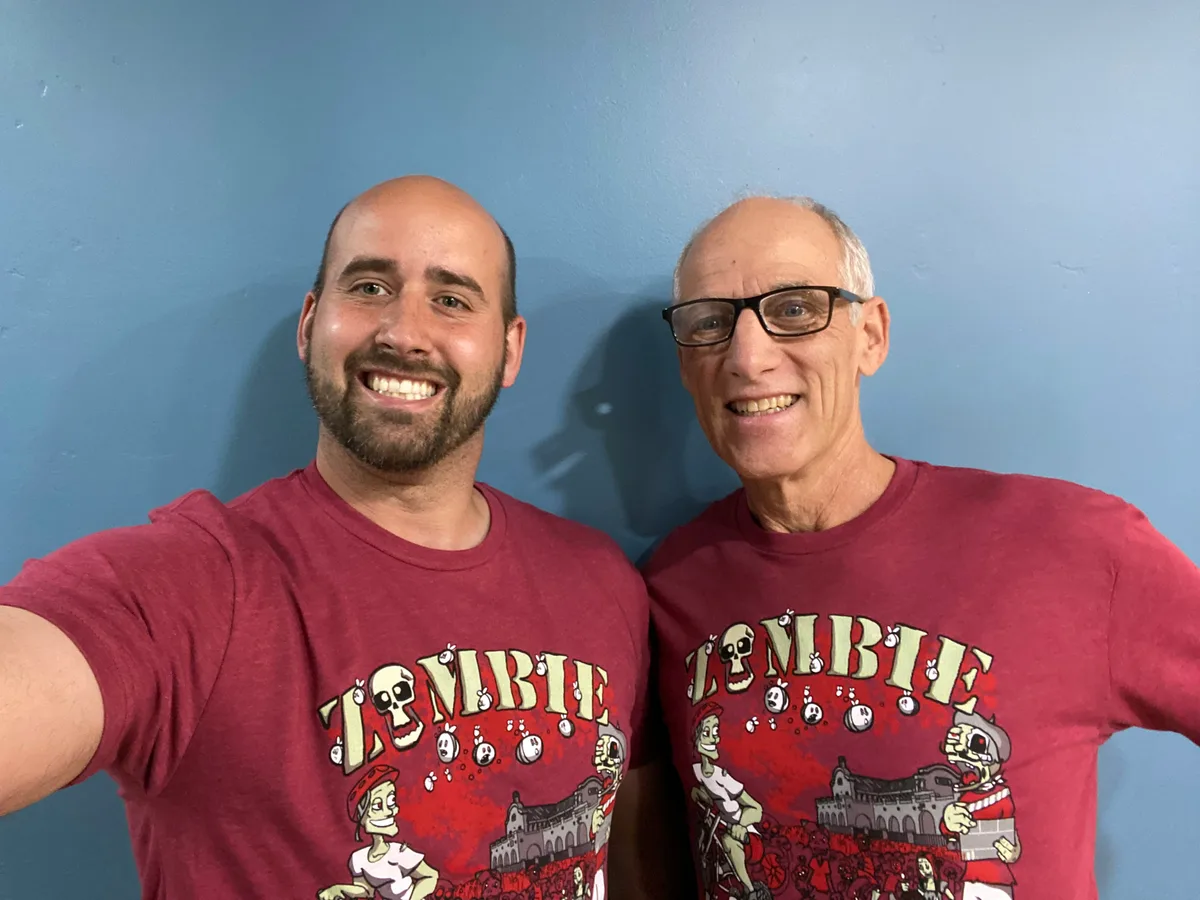Two men smile for a selfie wearing matching dark red Zombie Bike Parade t-shirts.