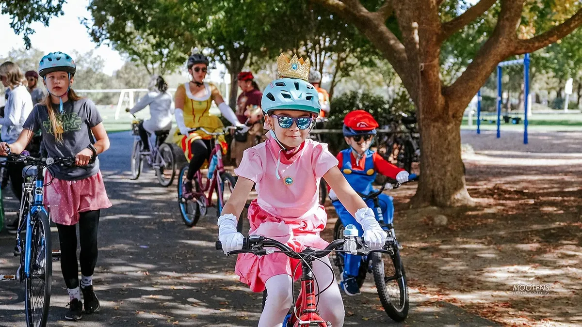 A young rider in a pink tutu and gold crown leads a group of costumed children on bikes along a tree-lined park path.