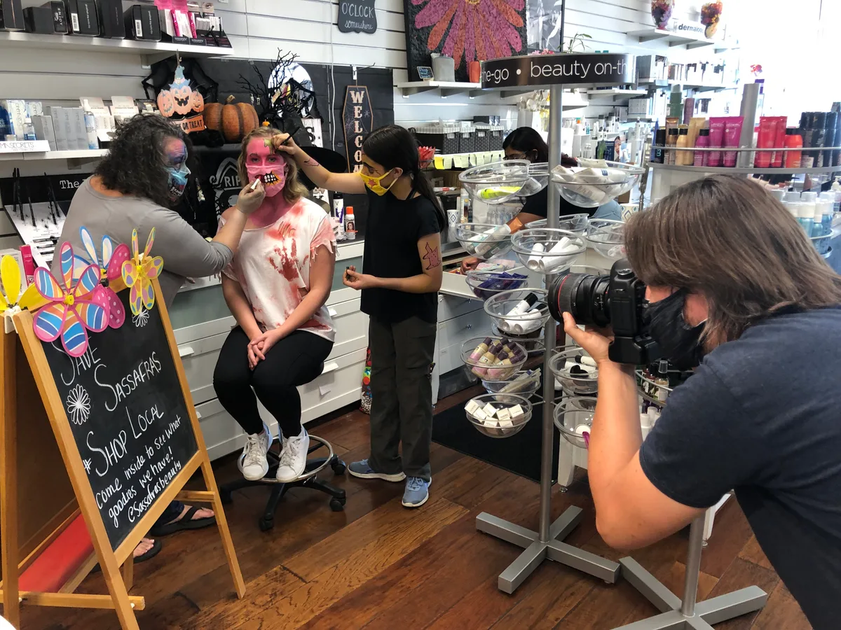 A photographer photographs a costumed woman at a beauty shop counter.