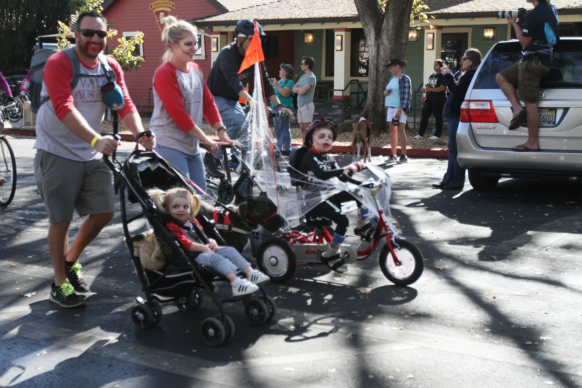 A family walks with a stroller near a red trike vehicle on a residential street.