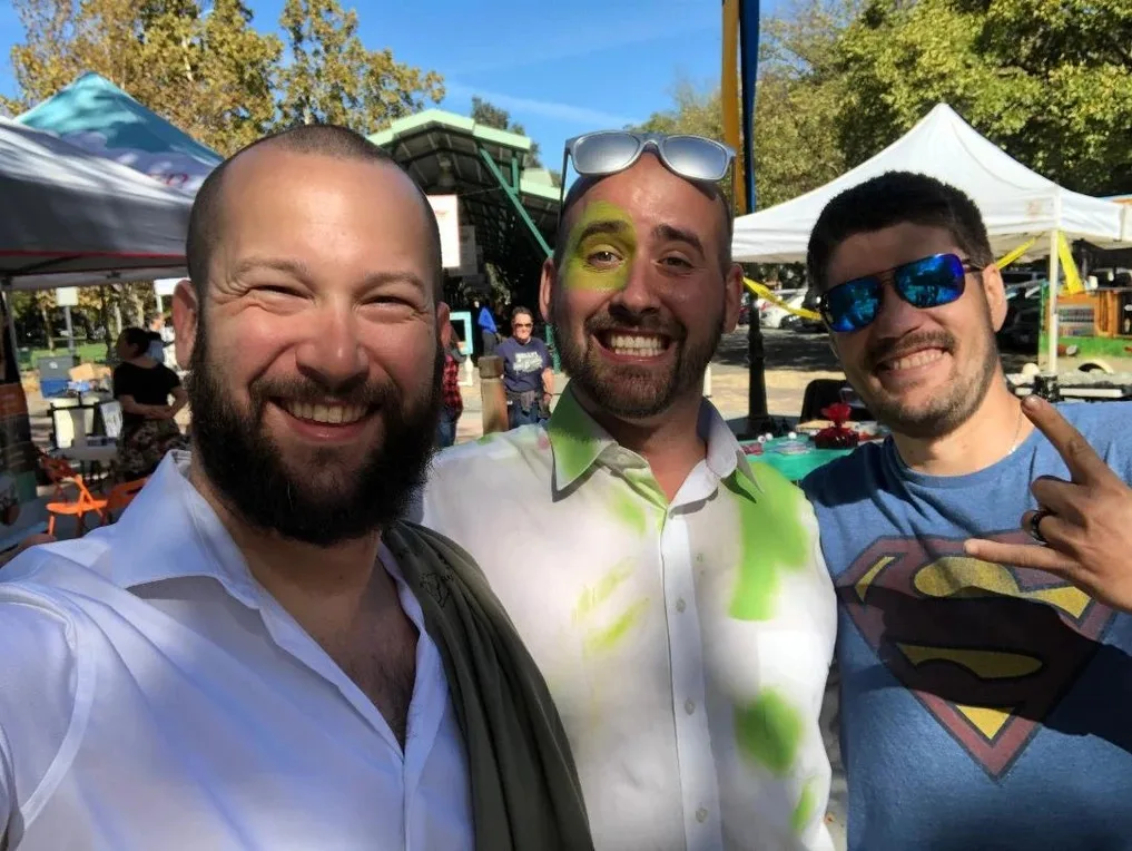 Three men pose together outdoors, one wearing a Superman shirt.
