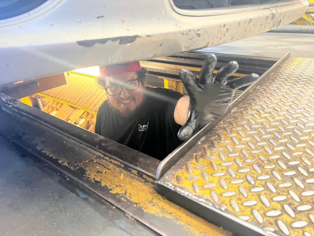 A grinning mechanic in zombie face paint waves up through the open service bay floor of a car lift.