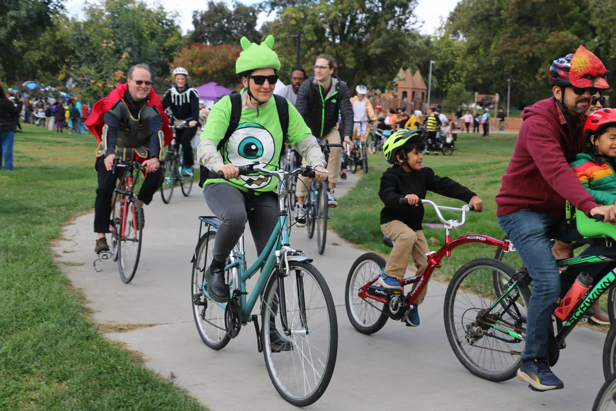 Riders in playful costume helmets pedal down the path with a young cyclist alongside.