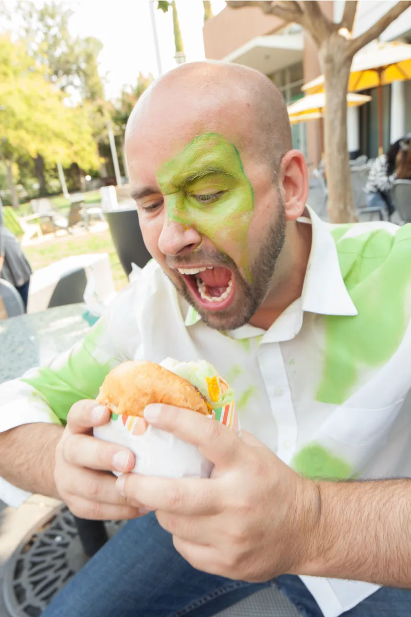 A man with green face paint opens his mouth wide in an exaggerated scream.