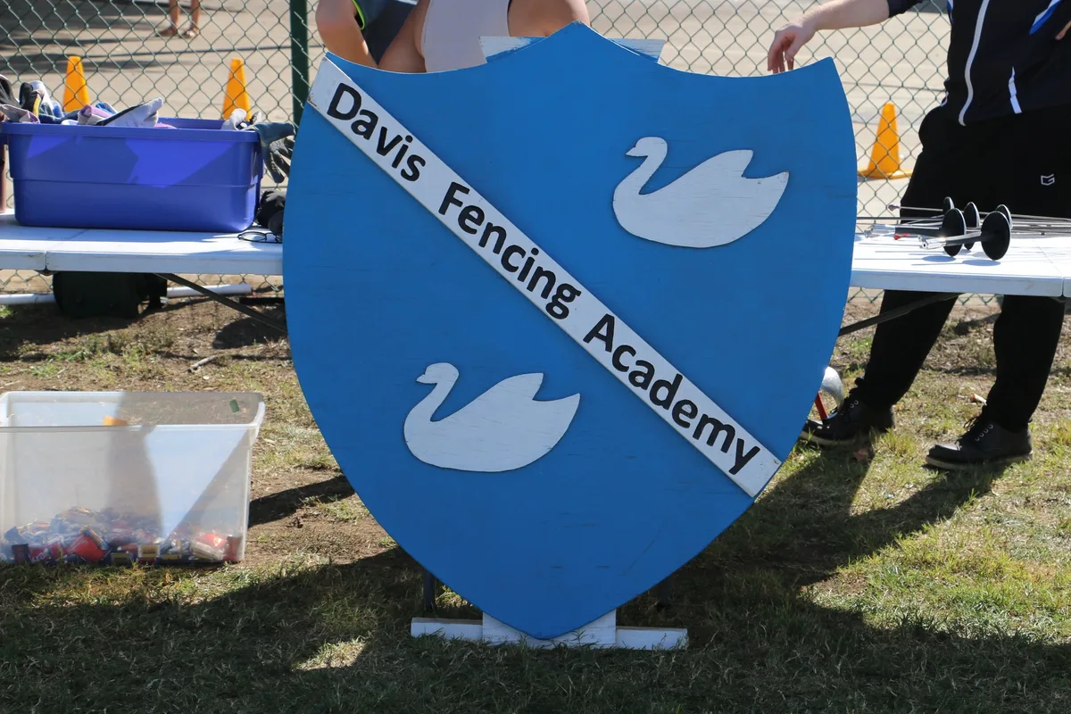 A Davis Fencing Academy sign stands in front of fencers practicing at a booth.