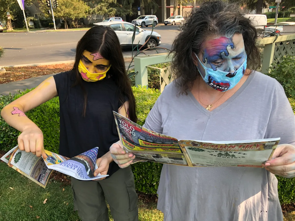 A woman and a girl in painted zombie face masks read copies of the Yolo County consumers guide newspaper on a residential street.