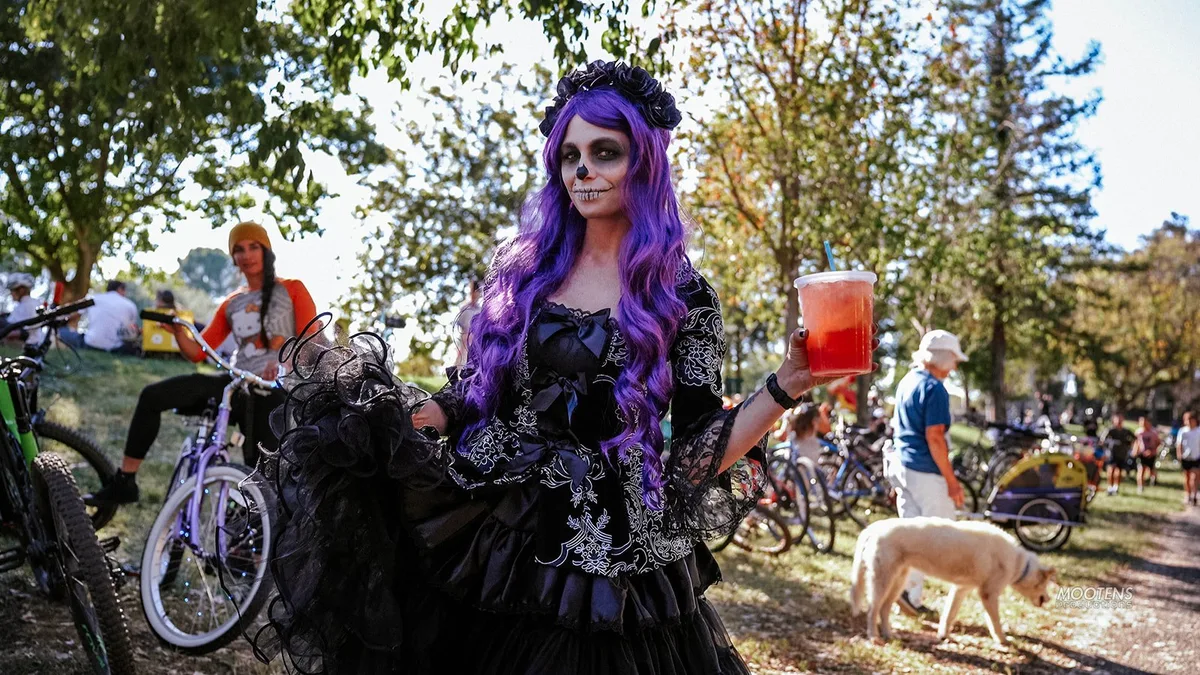 A woman in elaborate purple-haired Day of the Dead makeup and a black Victorian gown holds a drink in the park.