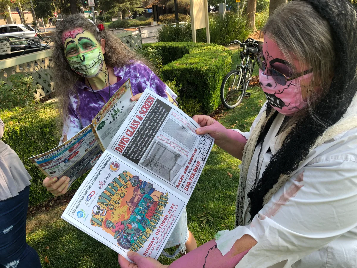 Two zombie-costumed women read a prop newspaper on the sidewalk.