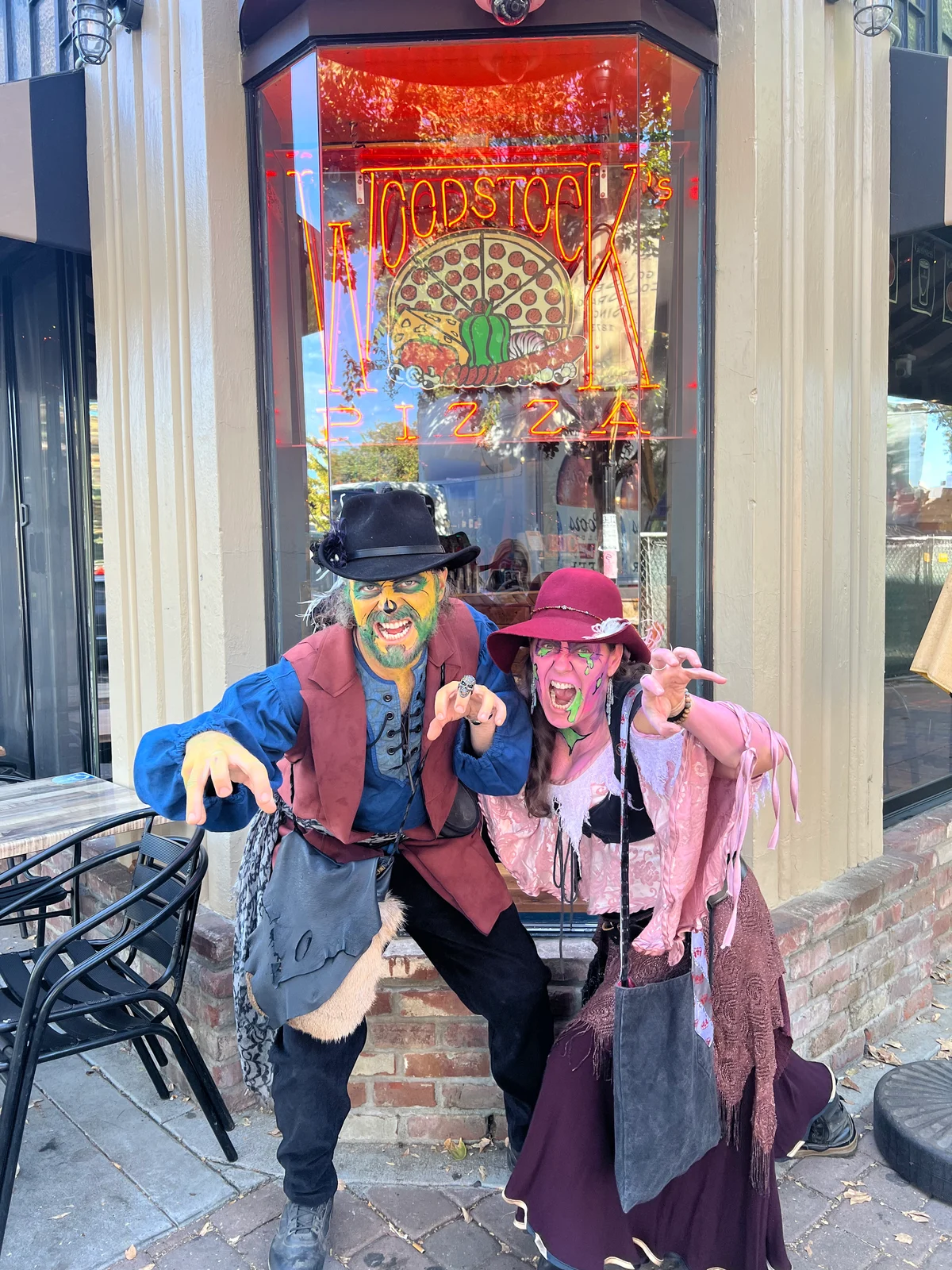 A zombie pirate and a zombie in a pink dress pose lurching outside Woodstock’s Pizza in downtown Davis, under the neon window sign.