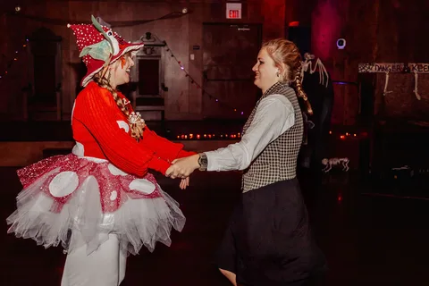 A woman in a mushroom costume dances hand in hand with a friend at the Zombie Ball, string lights glowing behind them.