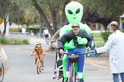 A laughing man in a giant inflatable alien costume rides his bike past other costumed riders on a park path.
