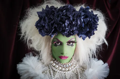A performer in green face makeup, a feathered headdress, and pearls poses in a dramatic studio portrait.