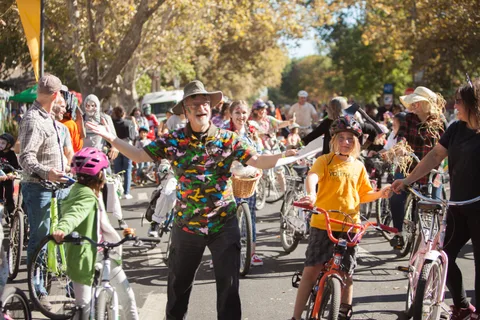 A costumed man greets a young rider as a huge crowd of cyclists fills a tree-lined street.