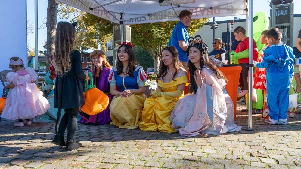 Four performers in Disney princess costumes sit outside the Davis Downtown booth on the Trick-or-Treat Trail, a pumpkin bucket beside them and children in costume queueing.