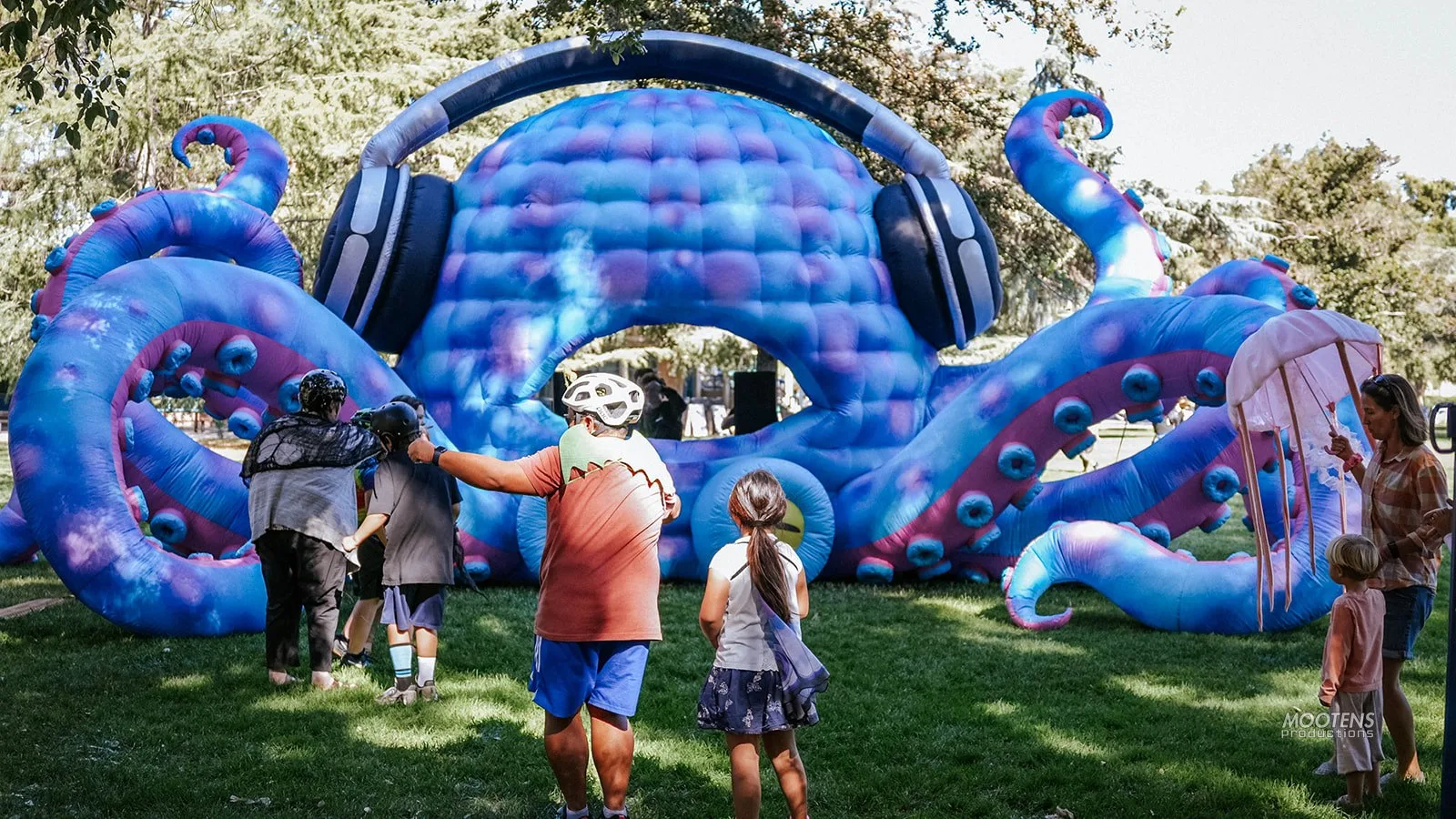 Kids and adults explore a giant inflatable octopus wearing oversized headphones in the park.