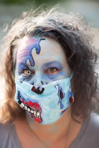 A close portrait of a woman with striking blue zombie face paint and a painted mask over a wide-eyed stare.