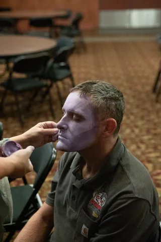 A man sits still with his eyes closed while a makeup artist sponges pale purple zombie makeup across his cheek, in a lodge hall set with round tables.