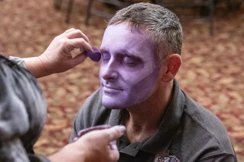 Close-up of a man having lavender zombie makeup blended around his eye with a small sponge, his eyes shut and a faint smile on his face.