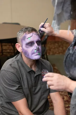 A makeup artist leans in with a fine brush to paint a black crack across the forehead of a man in purple and green zombie makeup.