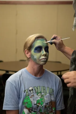A woman in a Zombie Bike Parade t-shirt has green zombie makeup with painted skull teeth finished off at her eyebrow with a brush.