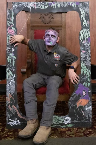 A man in purple and green zombie makeup sits in a carved wooden lodge chair, framed by a hand-painted board of cobwebs, bones and dancing zombies.